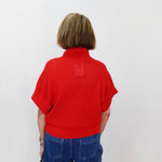 Person wearing a red sweater with a small logo on the back, standing against a white wall.