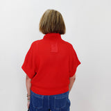 Person wearing a red sweater with a small logo on the back, standing against a white wall.