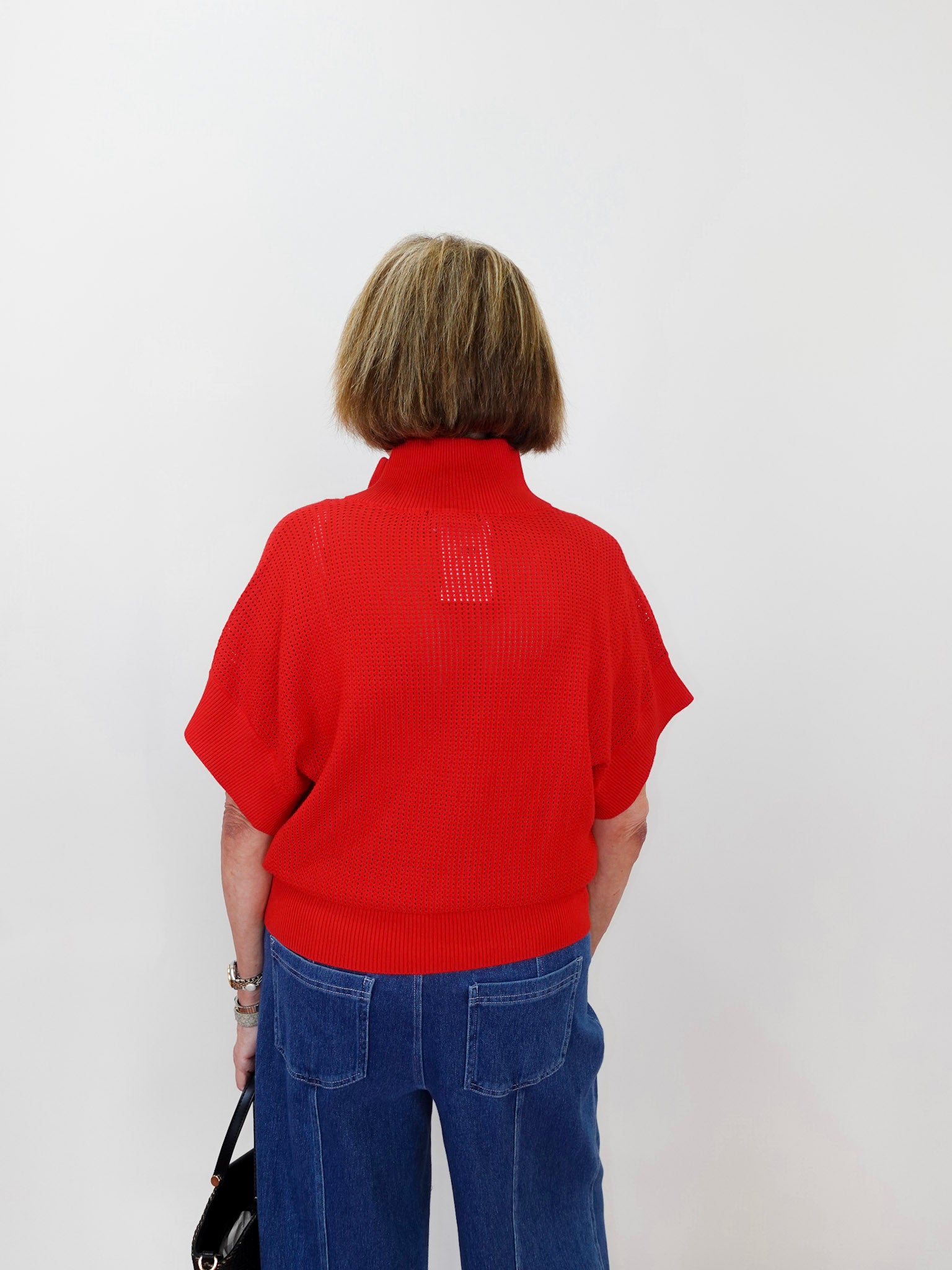 Person wearing a red sweater with a small logo on the back, standing against a white wall.