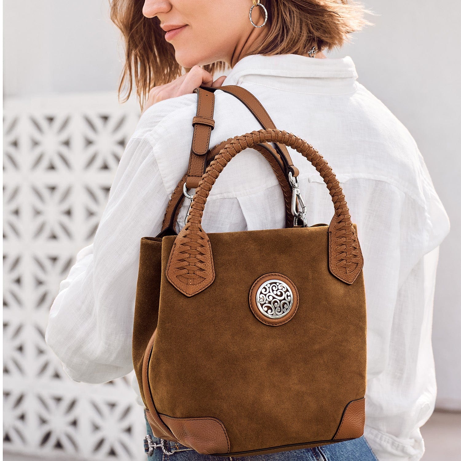 Brown suede handbag with leather handles and decorative emblem worn by a person against a white background
