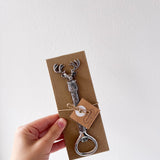Hand holding a deer-shaped bottle opener on a brown card with a white background