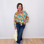 Woman wearing a colorful patterned shirt and jeans, holding a beige bag against a white wall.