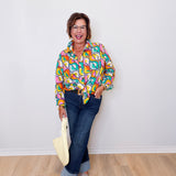 Woman wearing a colorful patterned shirt and jeans, holding a beige bag against a white wall.