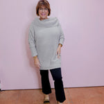 Woman wearing a light gray sweater and black pants against a pink wall.