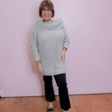 Woman wearing a light gray sweater and black pants against a pink wall.