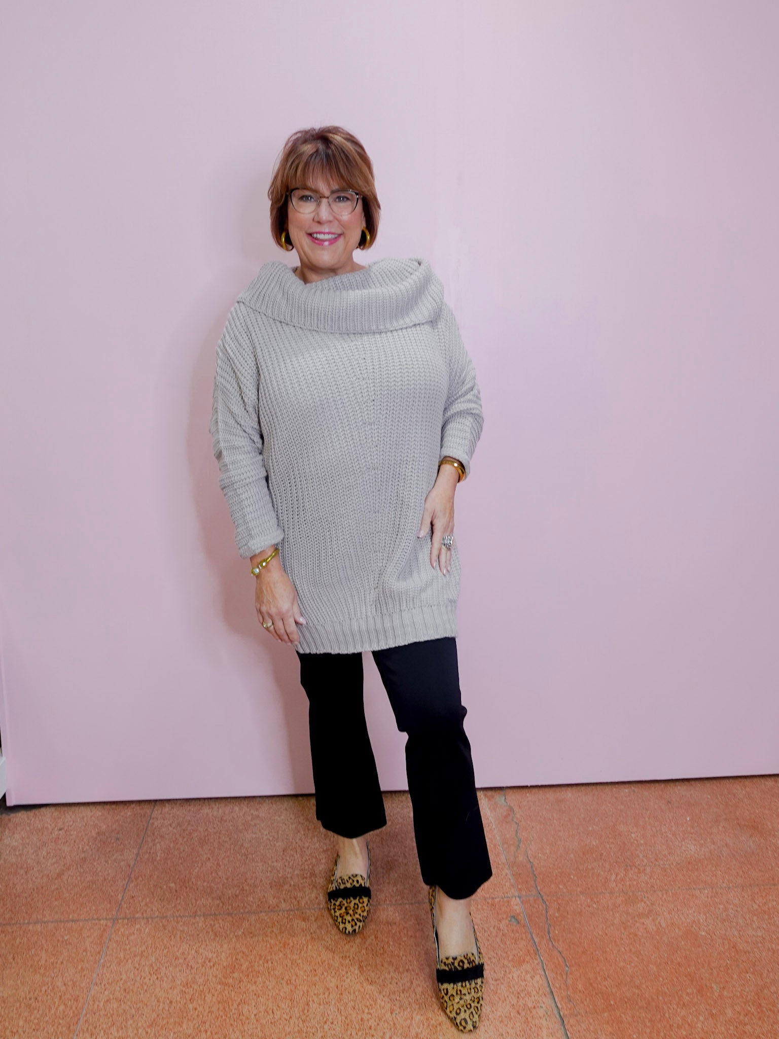 Woman wearing a light gray sweater and black pants against a pink wall.