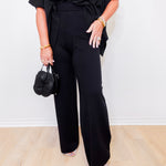Person wearing a black outfit with wide-leg pants holding a black handbag against a white background