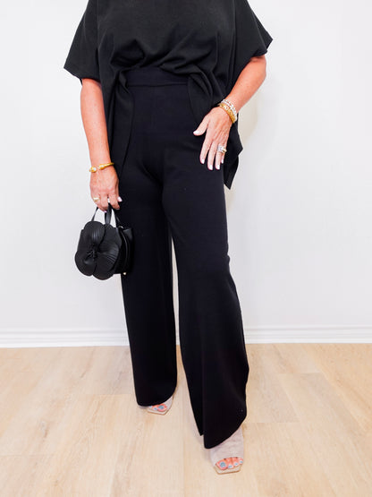 Person wearing a black outfit with wide-leg pants holding a black handbag against a white background
