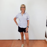 Woman wearing a light blue shirt, black shorts, and white sneakers standing against a white wall.