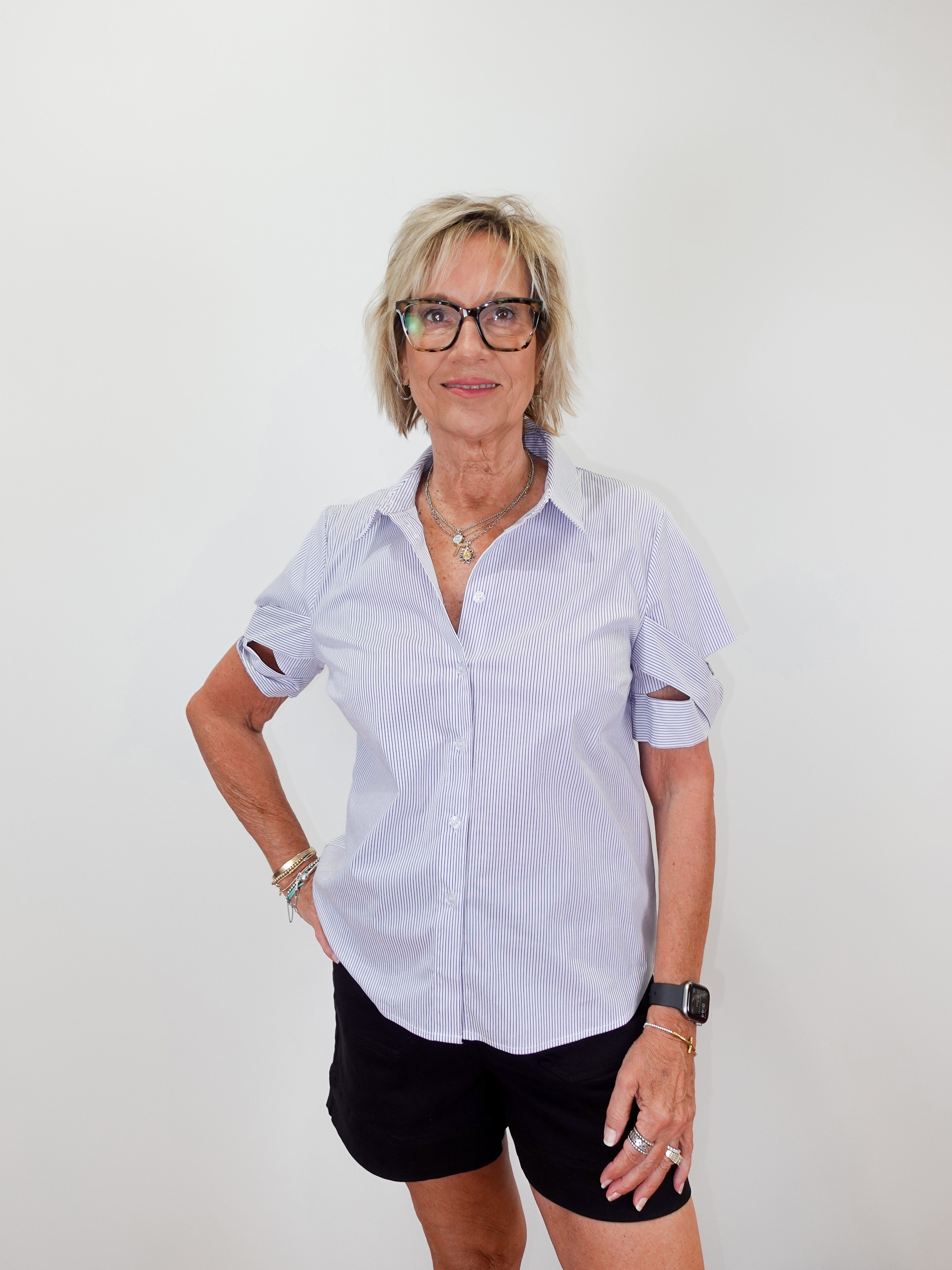 Woman wearing a light blue shirt, black shorts, and white sneakers standing against a white wall.