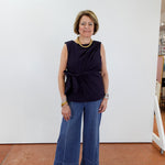 Woman wearing a navy sleeveless top and blue jeans standing against a white wall.