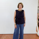 Woman wearing a navy sleeveless top and blue jeans standing against a white wall.
