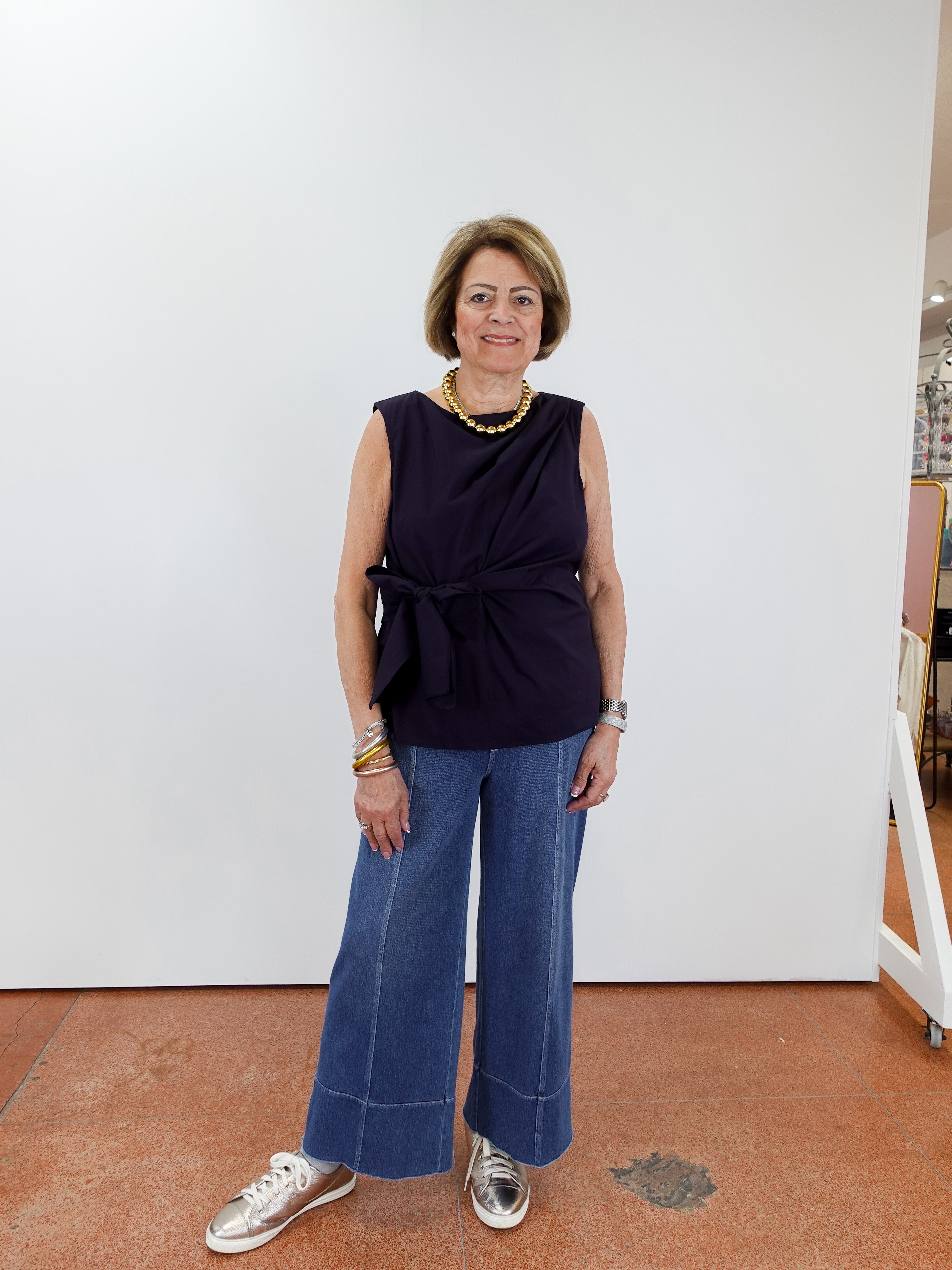 Woman wearing a navy sleeveless top and blue jeans standing against a white wall.