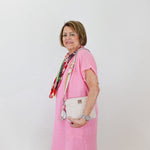 Woman wearing a pink dress with a colorful scarf and white handbag against a plain background