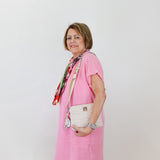 Woman wearing a pink dress with a colorful scarf and white handbag against a plain background