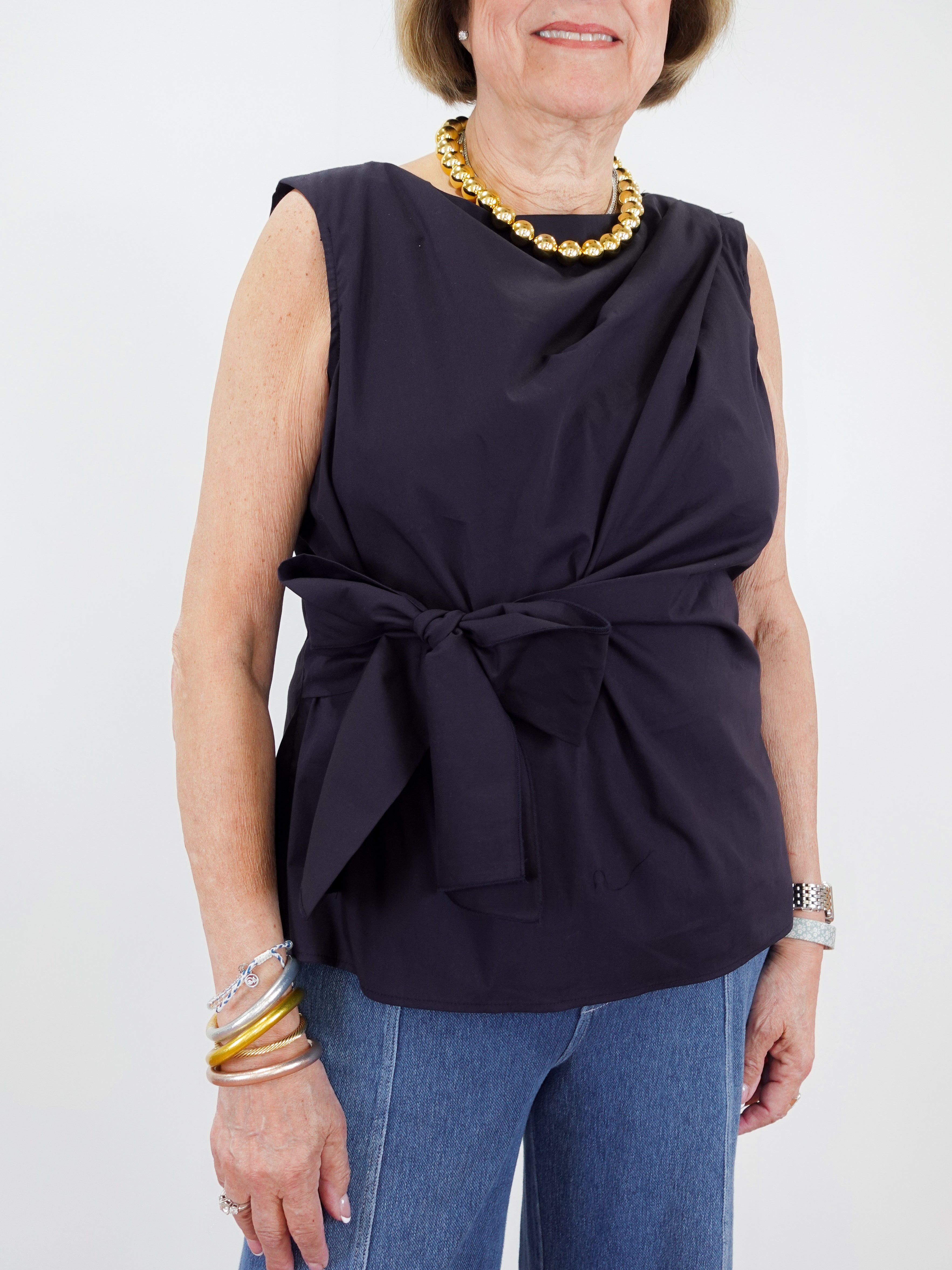 Woman wearing a navy sleeveless top with a bow detail and blue jeans on a white background