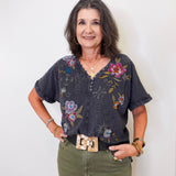 Woman wearing a floral blouse and green pants against a white wall