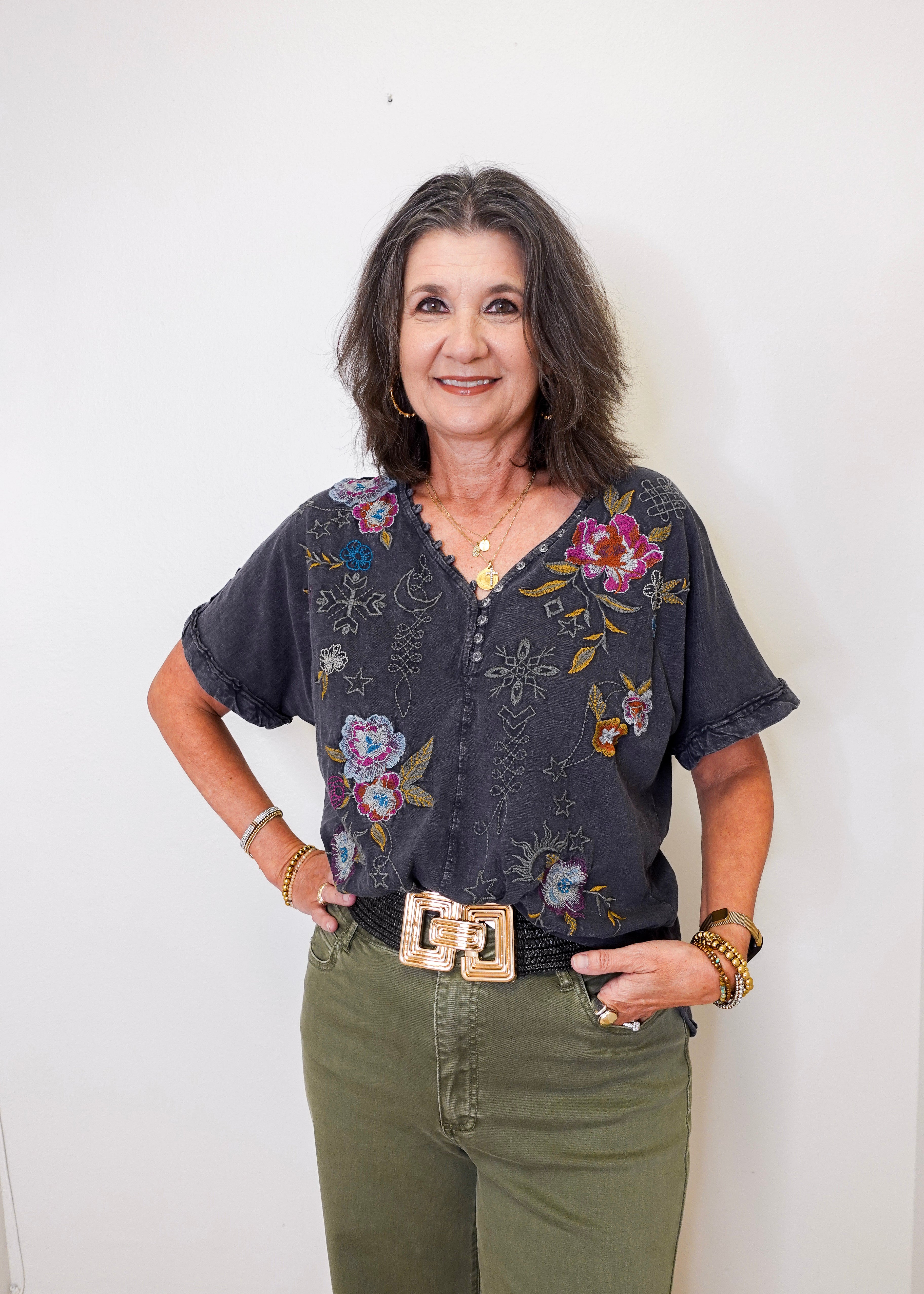 Woman wearing a floral blouse and green pants against a white wall