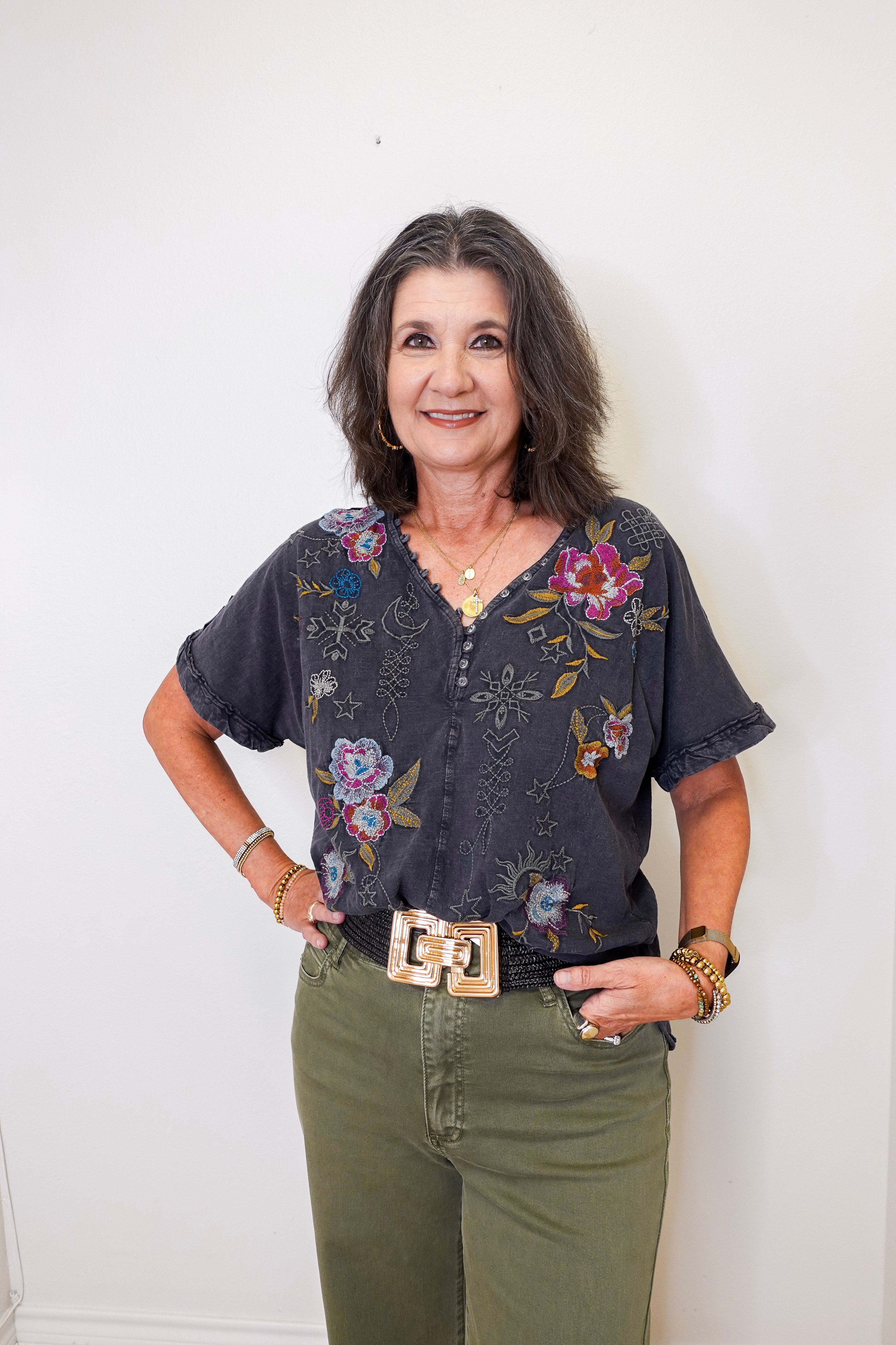 Woman wearing a floral blouse and green pants against a white wall