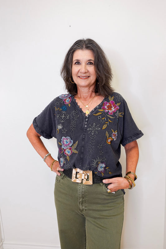Woman wearing a floral blouse and green pants against a white wall