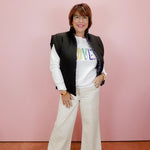 Woman wearing a black vest over a white shirt with colorful text, standing against a pink wall.