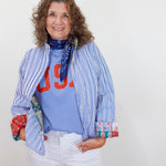 Woman wearing a blue striped jacket over a colorful shirt with text, white pants, and a blue scarf.