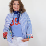 Woman wearing a blue striped jacket over a colorful shirt with text, white pants, and a blue scarf.