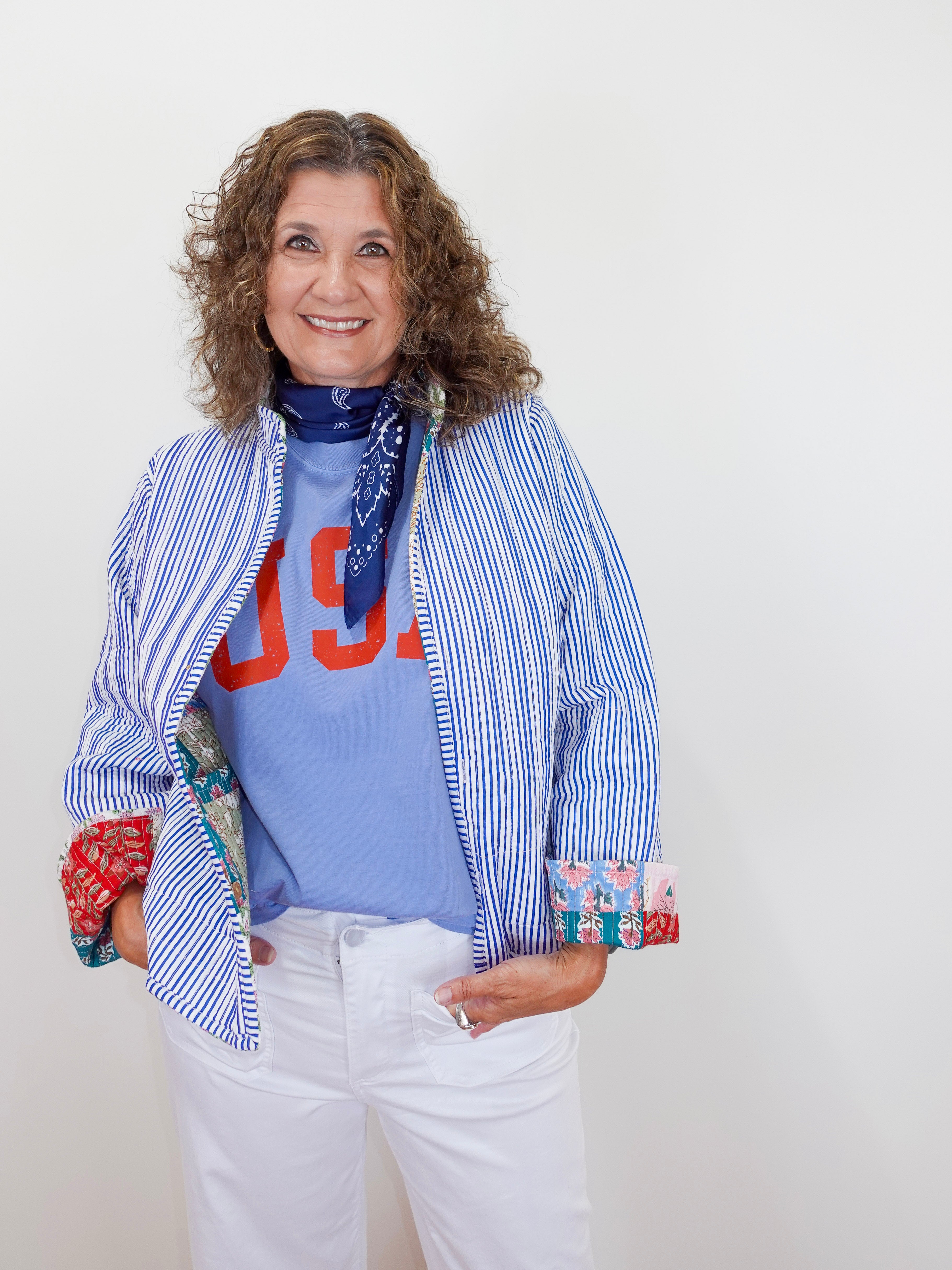 Woman wearing a blue striped jacket over a colorful shirt with text, white pants, and a blue scarf.