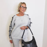 Woman wearing a gray outfit with black accents, holding a black handbag against a white wall.