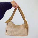 Beige woven handbag with a knotted handle held by a person wearing a dark blue sweater.
