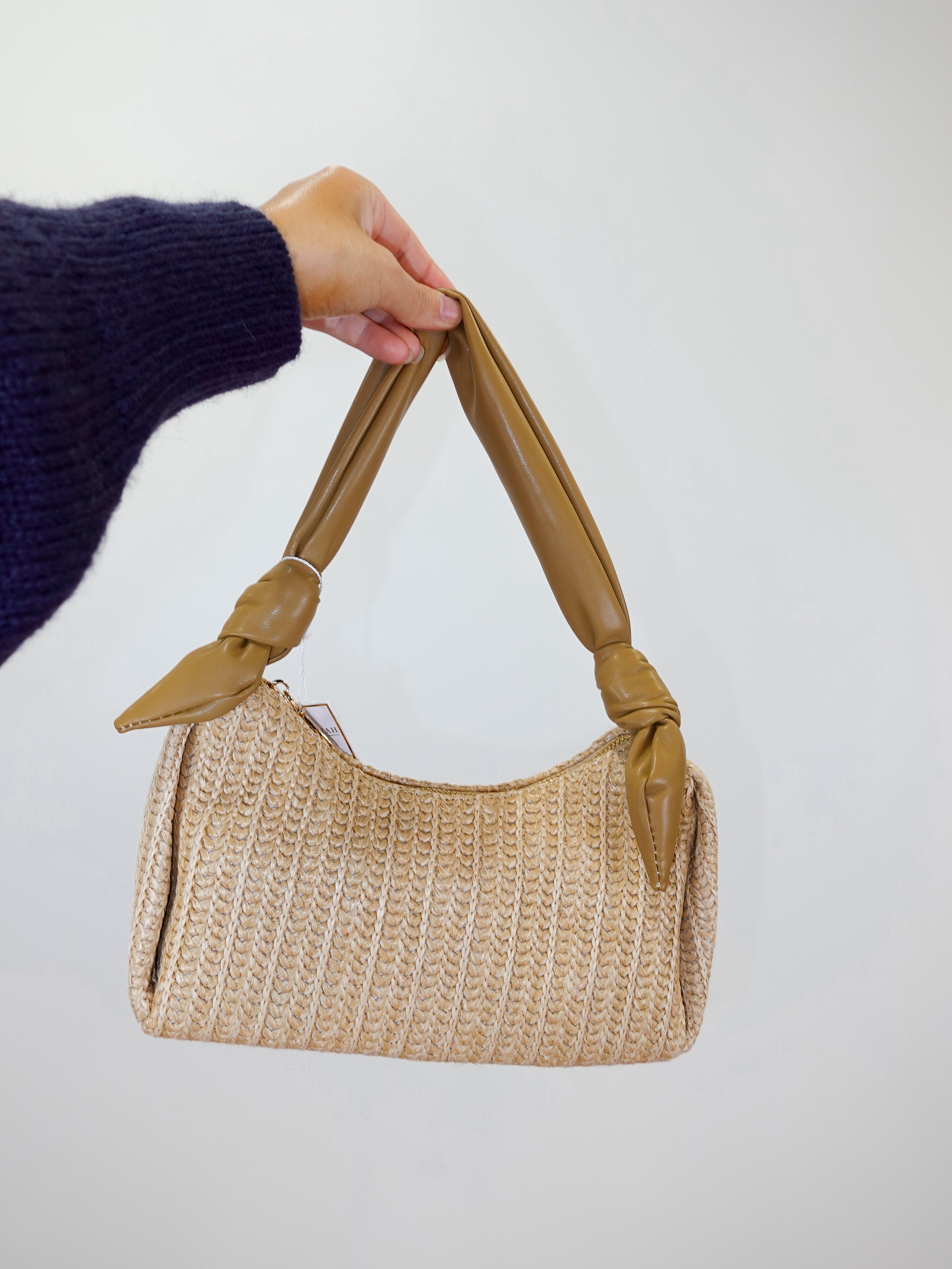 Beige woven handbag with a knotted handle held by a person wearing a dark blue sweater.