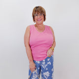Woman wearing a pink tank top and blue floral pants, holding a beige handbag against a white wall.