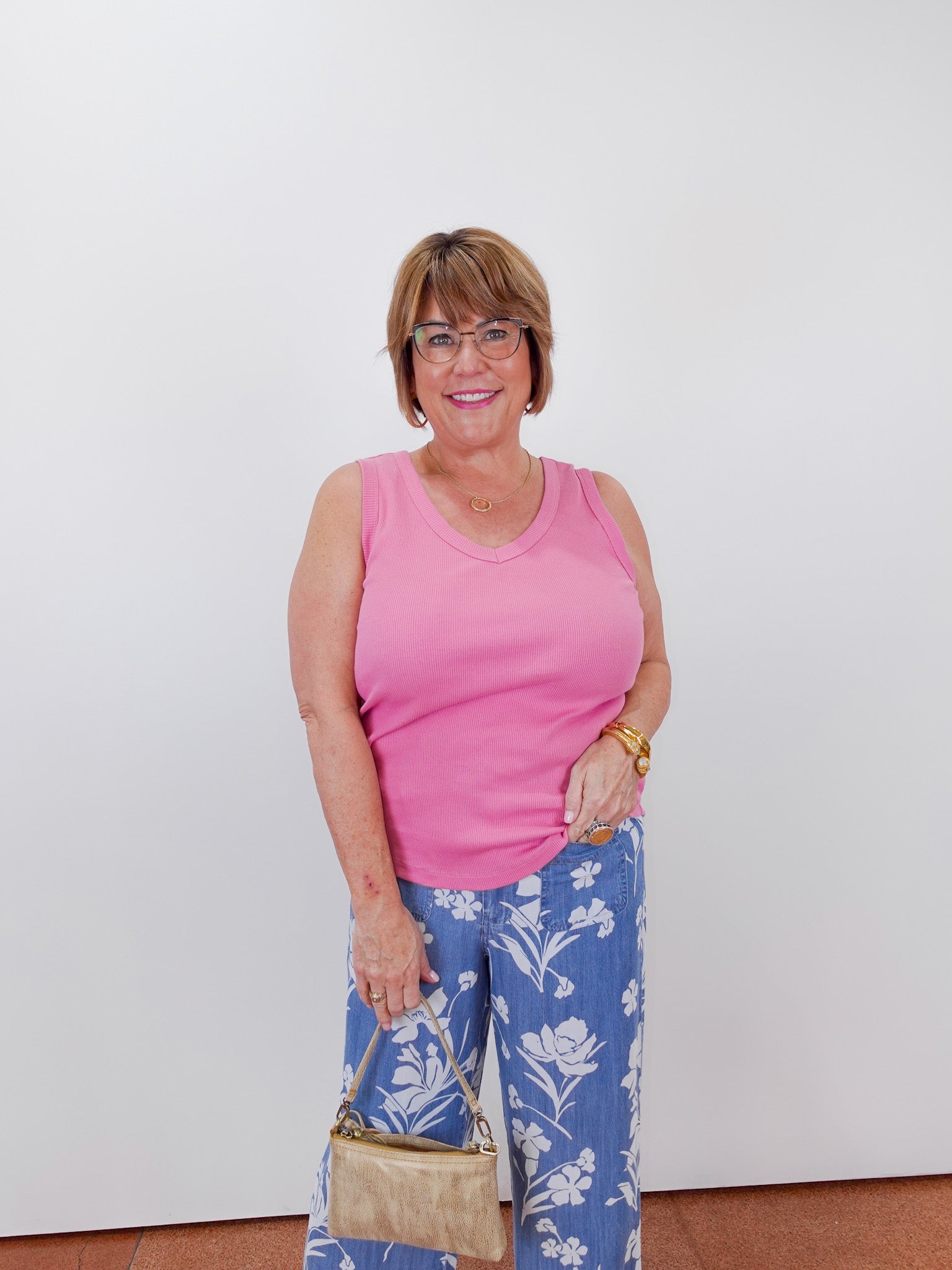 Woman wearing a pink tank top and blue floral pants, holding a beige handbag against a white wall.