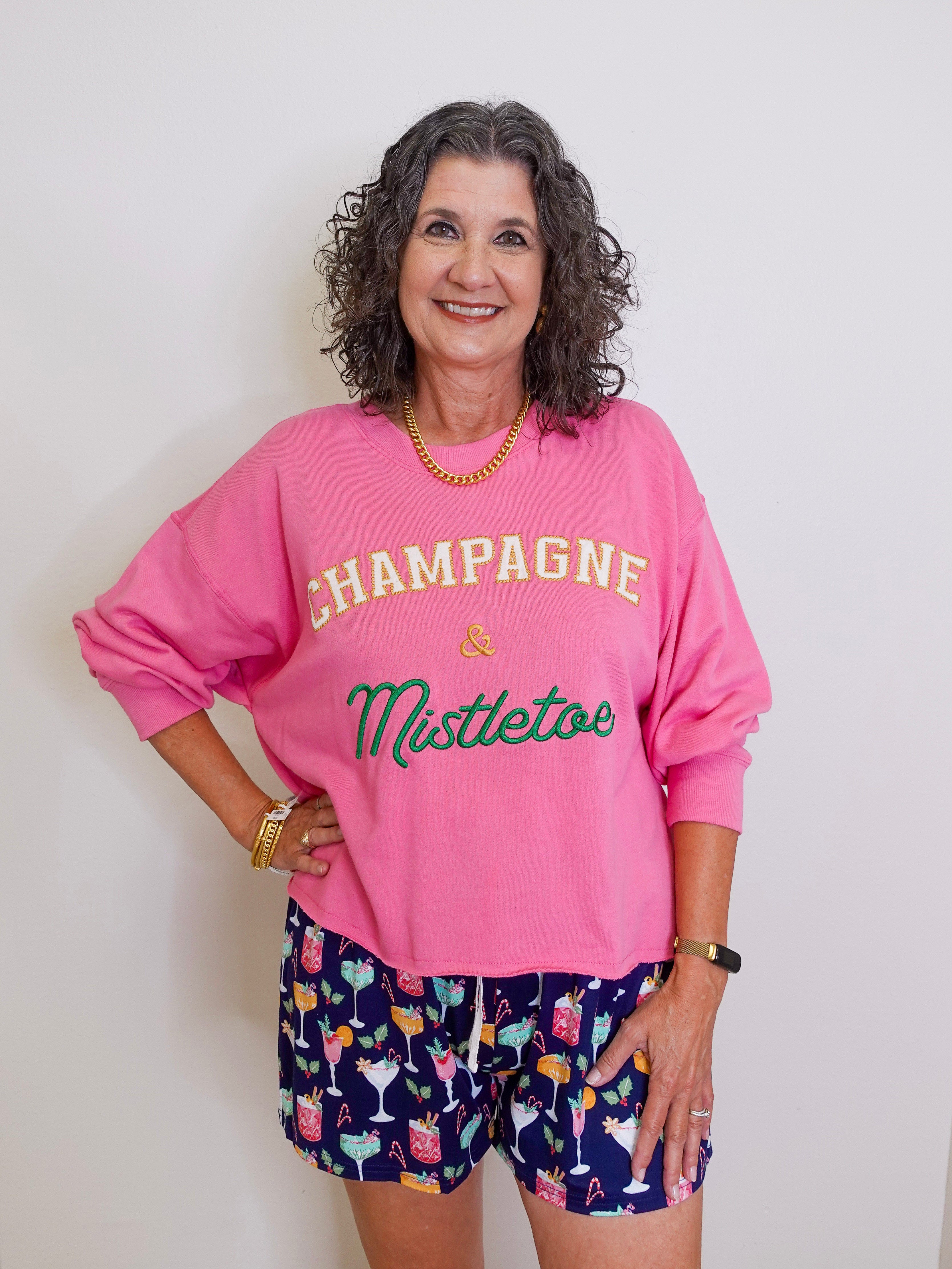 Woman wearing a pink sweatshirt with 'Champagne & Mistletoe' text and patterned shorts on a white background