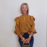 Woman wearing a mustard yellow blouse with ruffled sleeves against a white wall.