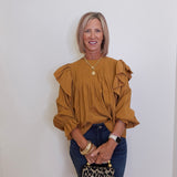 Woman wearing a mustard yellow blouse with ruffled sleeves against a white wall.