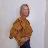 Woman wearing a mustard yellow blouse with ruffled sleeves against a white wall