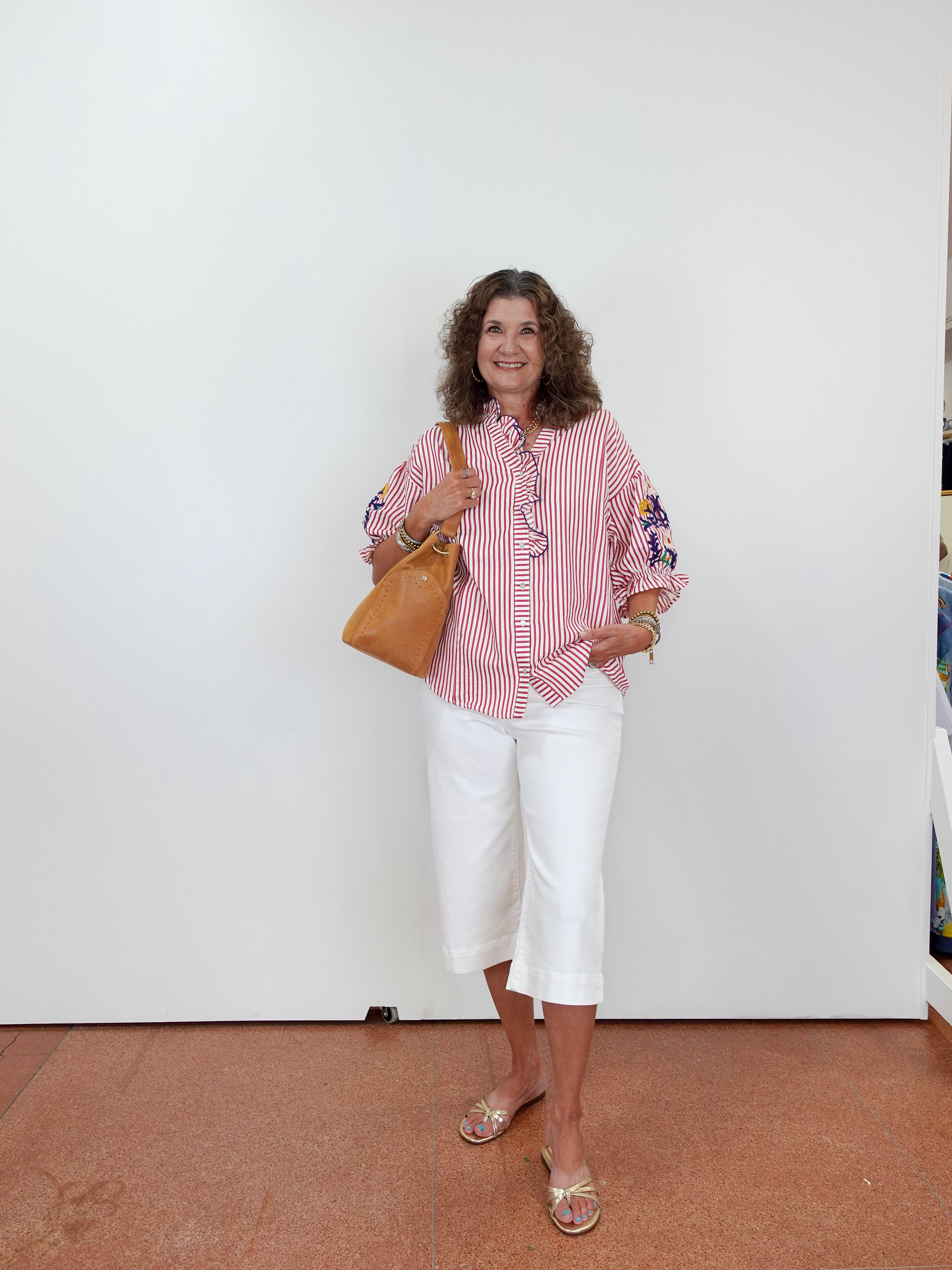 Woman wearing a red and white striped shirt with white pants, holding a brown bag against a white wall.