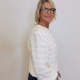 Woman wearing a white textured sweater and dark pants against a plain background
