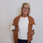 Woman wearing a brown jacket over a white sweater and dark pants against a plain background