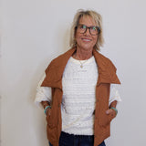 Woman wearing a brown jacket over a white sweater and dark pants against a plain background