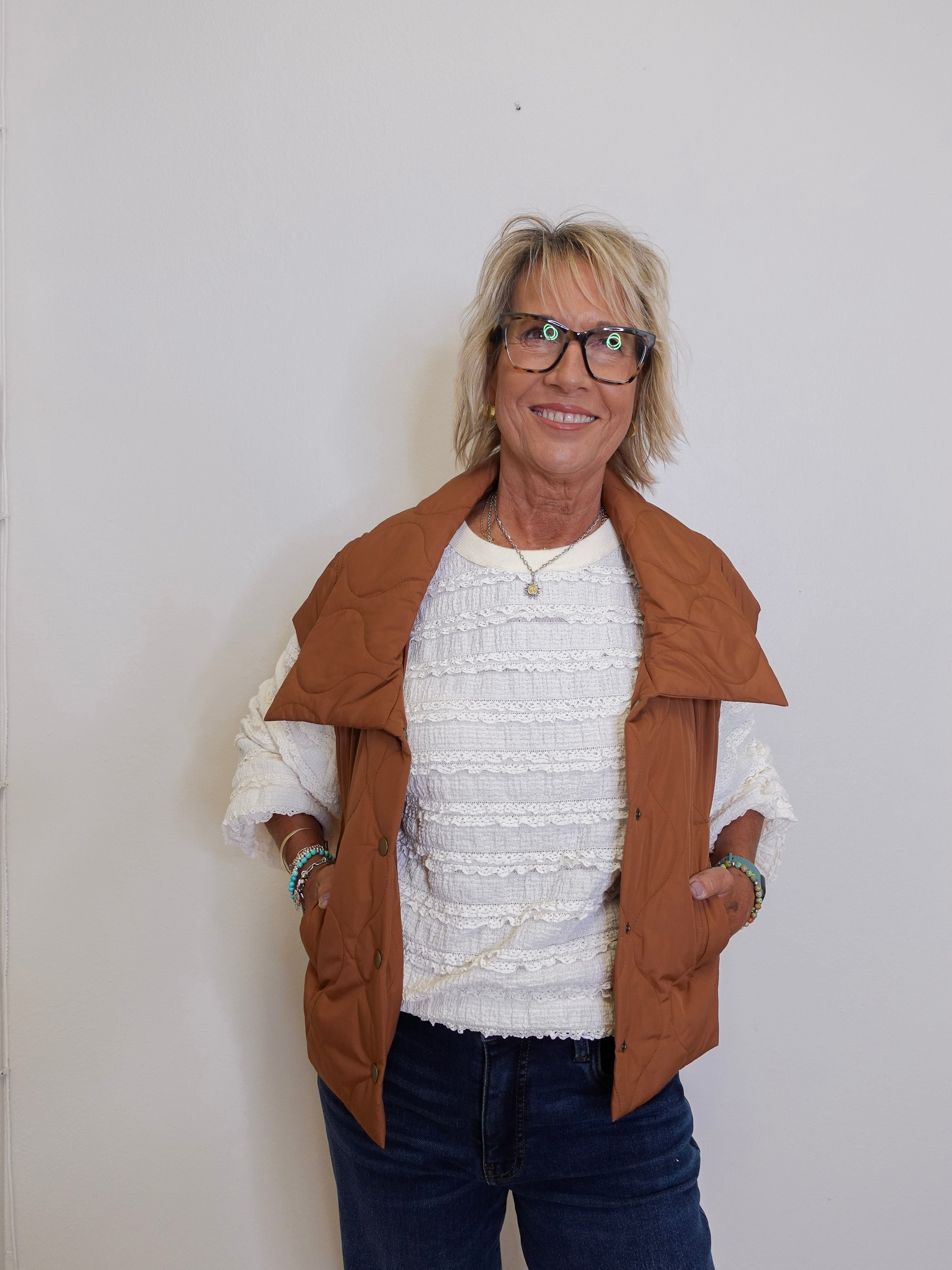 Woman wearing a brown jacket over a white sweater and dark pants against a plain background