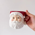 Hand holding a ceramic mug shaped like a Santa Claus face against a plain background