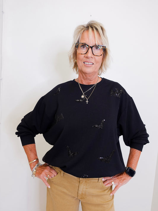 woman wearing a black sequin bat sweatshirt against a white wall. 