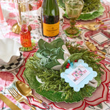 Decorative table setting with green wreath, wine bottle, and place card on a patterned tablecloth.