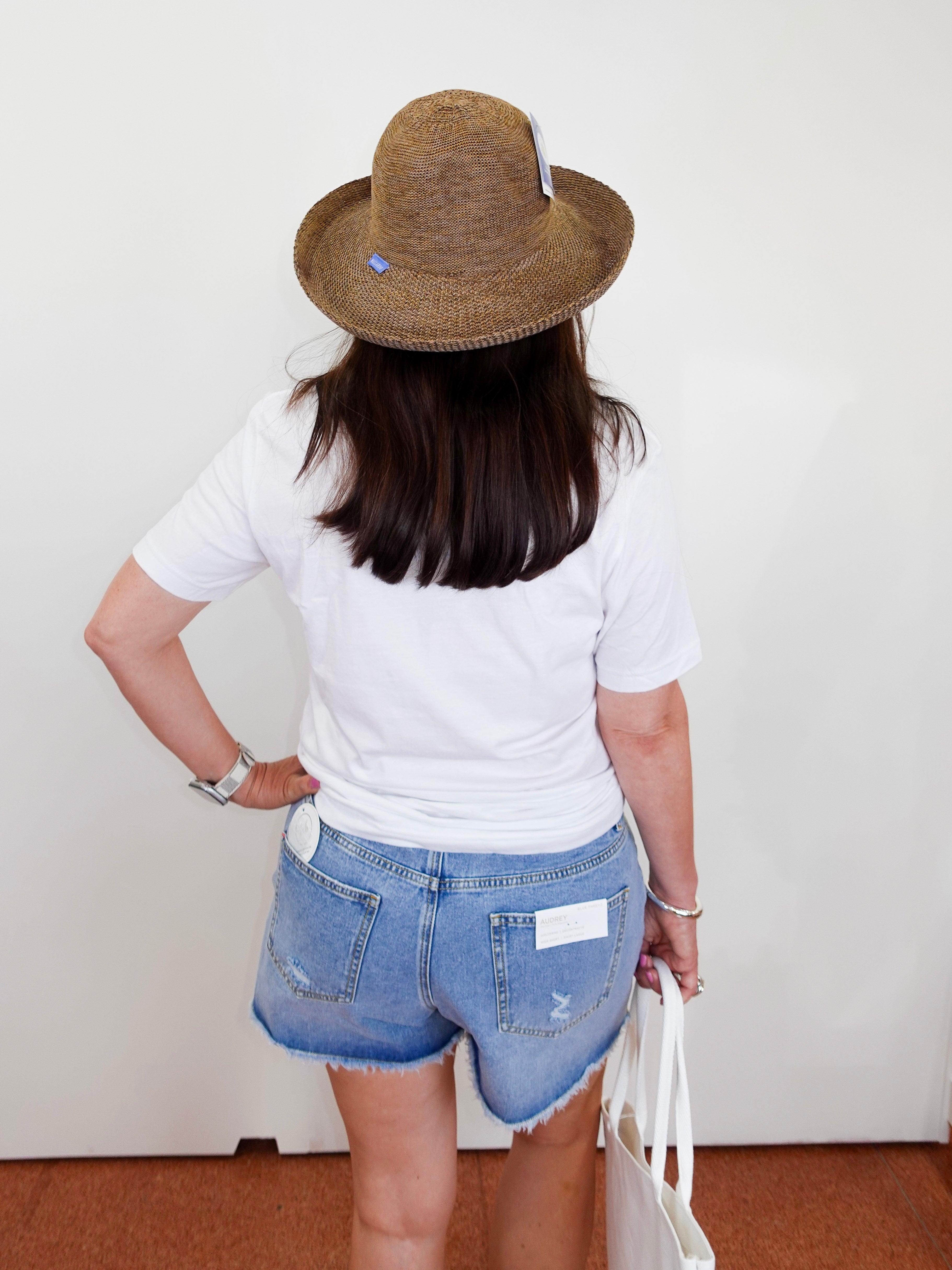Person wearing a straw hat, white t-shirt, and denim shorts against a plain background