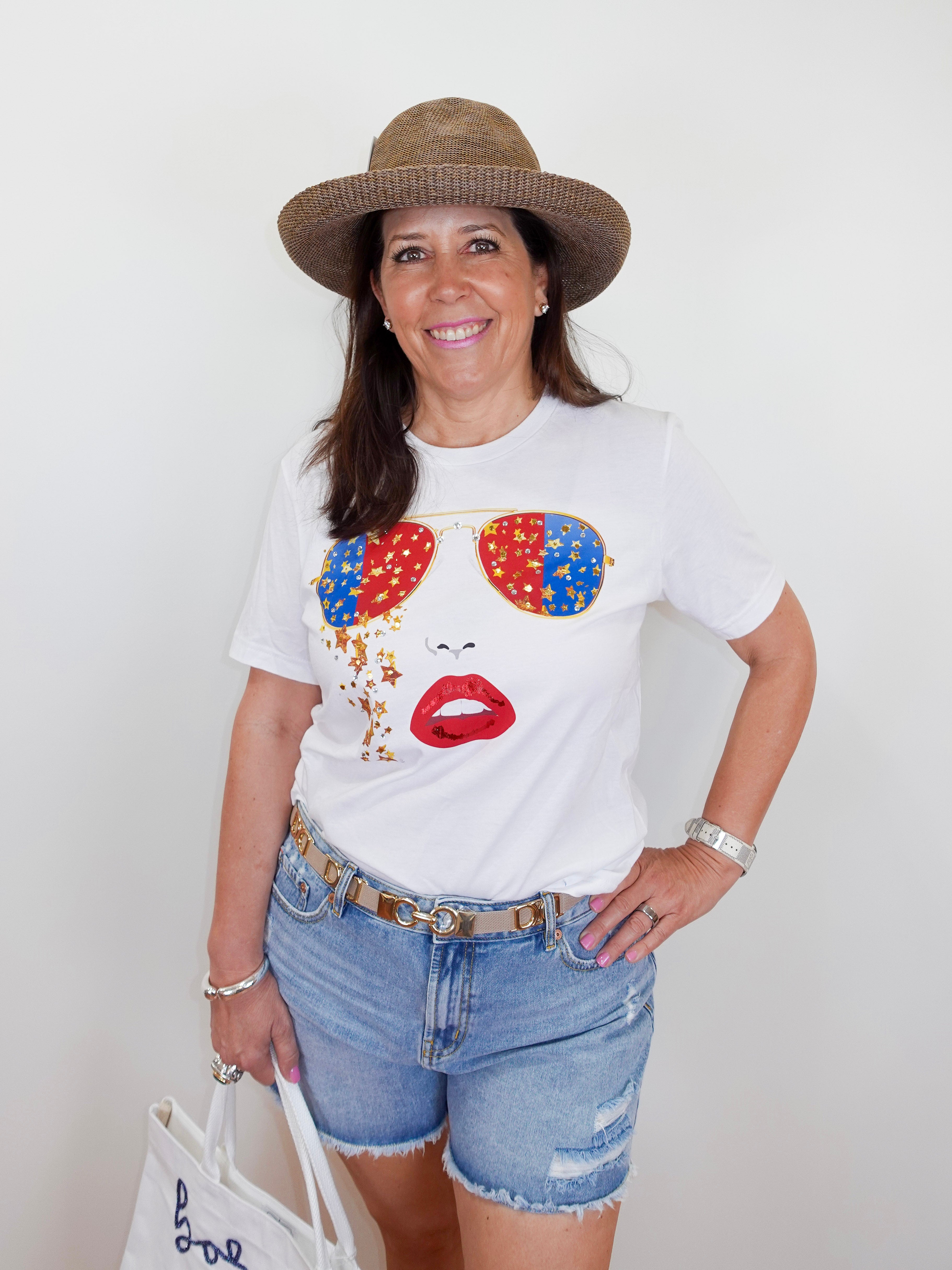 Woman wearing a white t-shirt with colorful sunglasses and lips design, denim shorts, and a straw hat.
