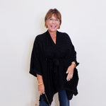 Woman wearing a black poncho and holding a patterned handbag against a white wall.