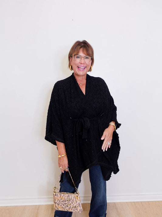 Woman wearing a black poncho and holding a patterned handbag against a white wall.