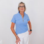 Woman wearing a light blue polo shirt and white pants against a plain background